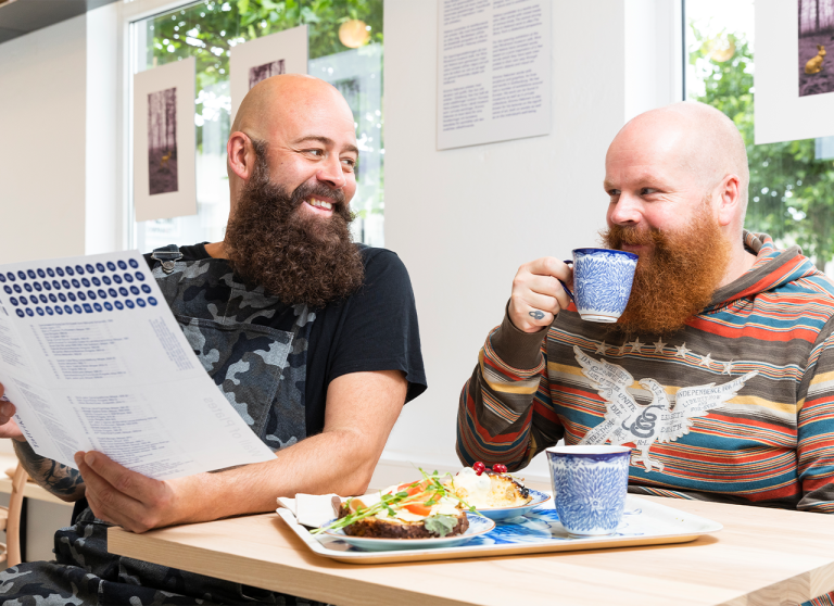 Två män i kafét tittar på varandra, den ena dricker kaffeo ch den andra tittar i häftet om tallriksväggen.