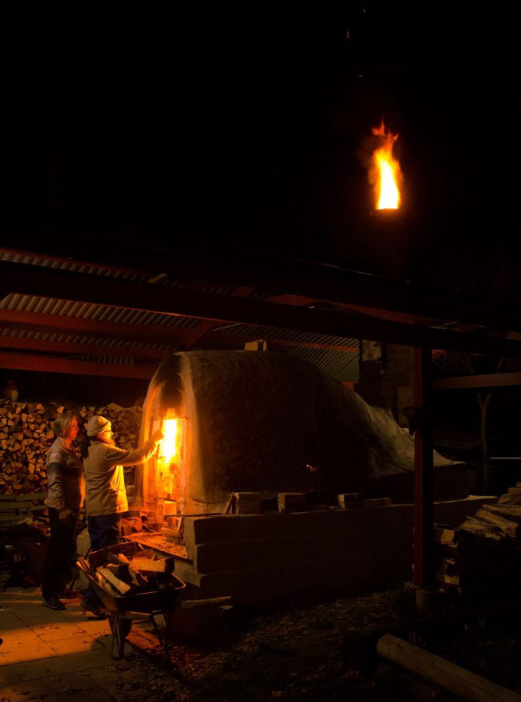 Två kvinnor intill en keramisk ugn där eld kommer ur skorstenen.
